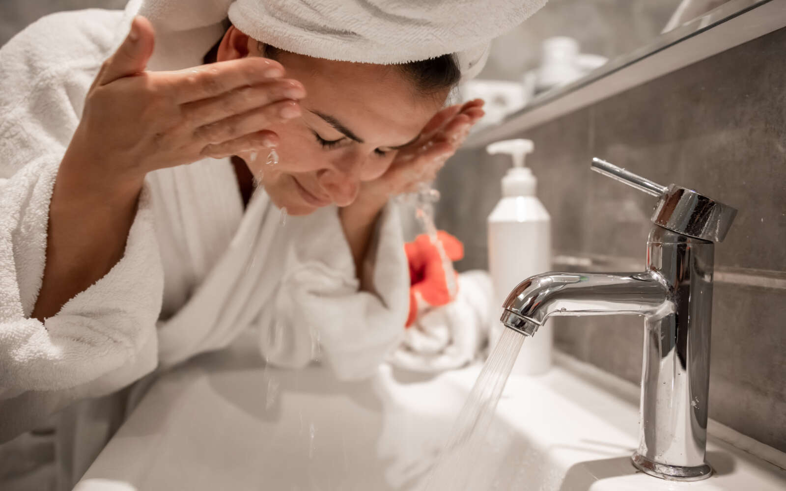 Mujer hermosa joven en el baño con una toalla en la cabeza se lava la cara con agua del grifo