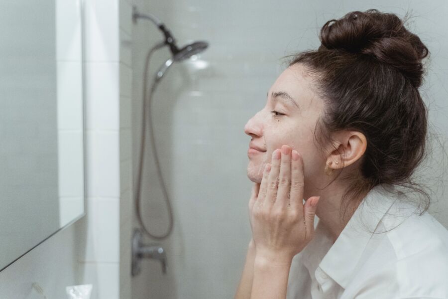 Una mujer aplica crema facial en un espejo para su rutina de cuidado de la piel en un baño.