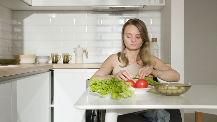 Mujer sentada a la mesa preparando verduras frescas en una cocina moderna, destacando la vida saludable y la independencia.