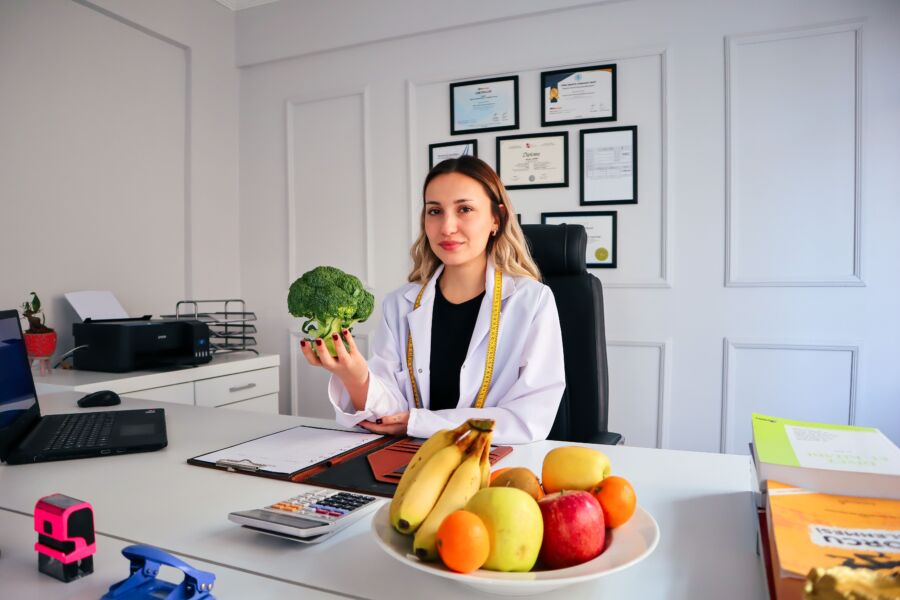 Nutricionista en oficina sosteniendo brócoli, rodeada de frutas, promoviendo un estilo de vida saludable.