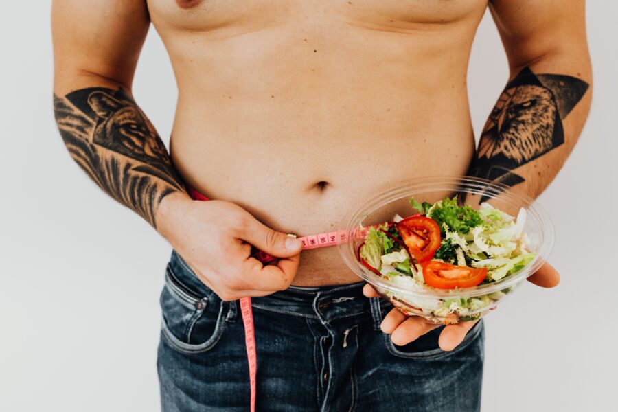 Hombre tatuado midiendo la cintura mientras sostiene ensalada fresca, simbolizando un estilo de vida saludable y control de peso.