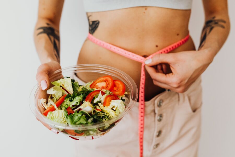 Mujer sosteniendo ensalada de verduras y midiendo la cintura con cinta adhesiva, simbolizando opciones de estilo de vida saludables.