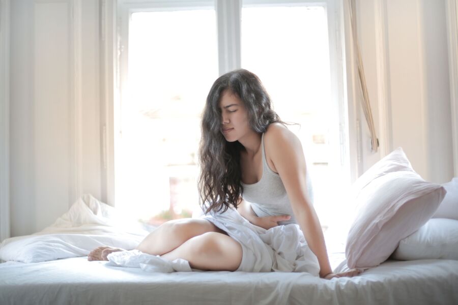 Mujer sentada en la cama sosteniéndose el estómago, mostrando signos de incomodidad o dolor en el interior.