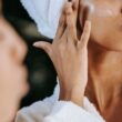 Recorte mujer étnica joven irreconocible en bata de rizo aplicando crema hidratante en la mejilla mientras se refleja en el espejo