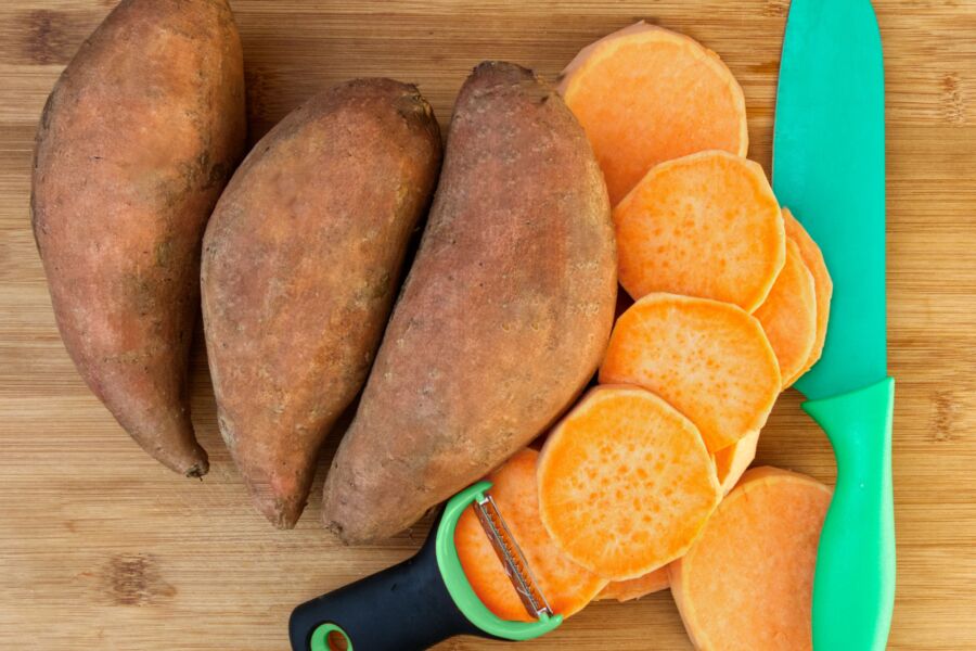 Primer plano de batatas, cortadas en rodajas con un cuchillo verde y peladora sobre una superficie de madera.