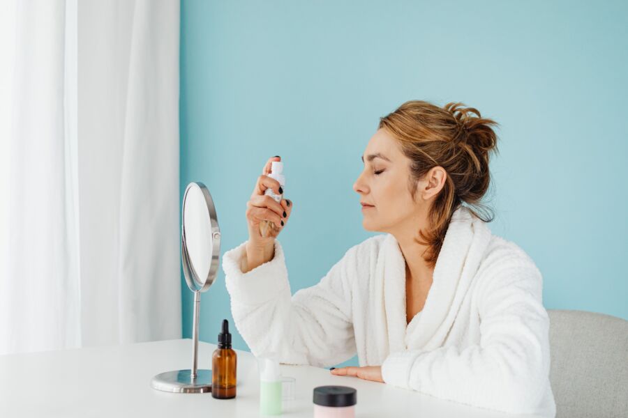 Mujer en bata de baño usando un spray para el cuidado de la piel frente a un espejo en el interior.