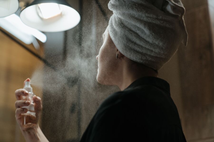 Mujer con una toalla en la cabeza aplicando spray facial en el baño, centrándose en el cuidado de la piel y el autocuidado.