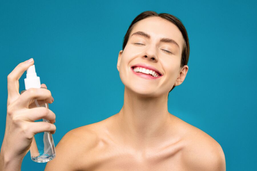 Una mujer alegre rocía la niebla facial con los ojos cerrados sobre un fondo azul, mostrando el cuidado de la piel y la felicidad.