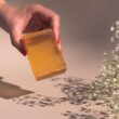 Una mano con esmalte de uñas rojo sostiene una barra de jabón naranja sobre las flores del aliento del bebé.
