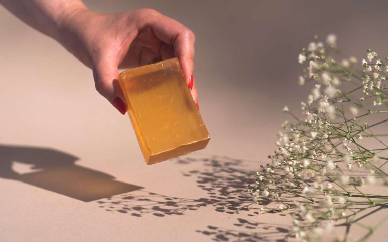 Una mano con esmalte de uñas rojo sostiene una barra de jabón naranja sobre las flores del aliento del bebé.