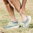 Hombre sosteniendo su pantorrilla con dolor, usando zapatillas de color verde azulado sobre hierba verde.