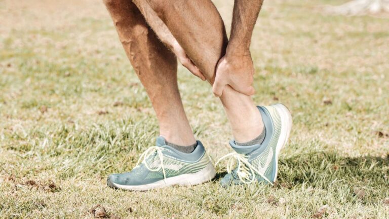 Hombre sosteniendo su pantorrilla con dolor, usando zapatillas de color verde azulado sobre hierba verde.