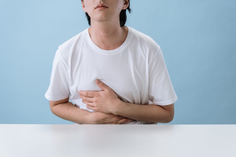 Un hombre adulto con una camisa blanca sostiene su estómago, lo que indica dolor e incomodidad, con un fondo azul claro.