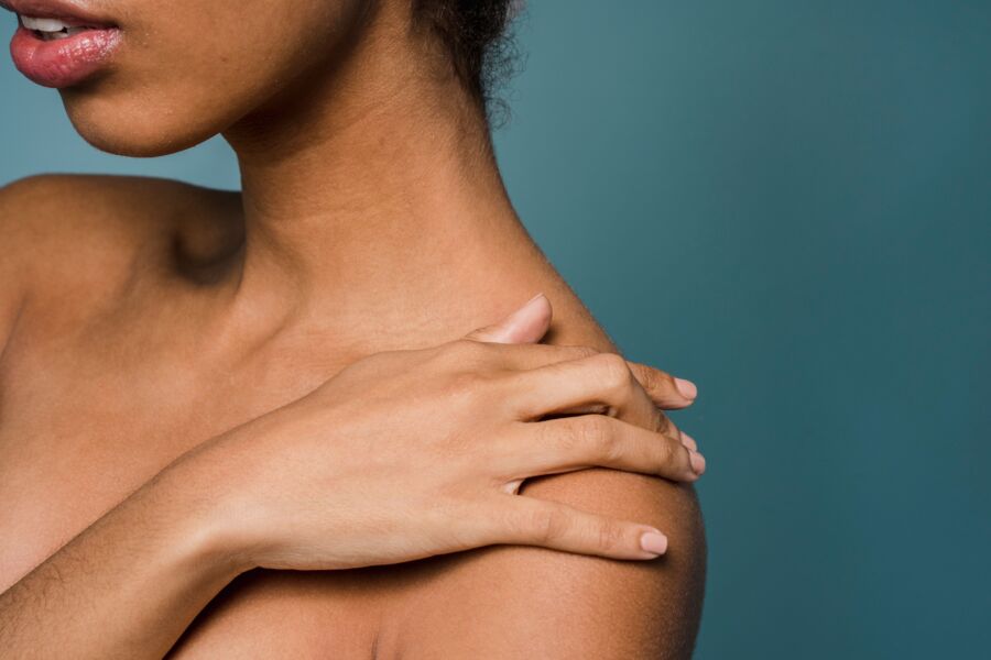 Vista lateral de una joven mujer negra agraciada y anónima tocando el hombro desnudo mientras está de pie sobre un fondo azul