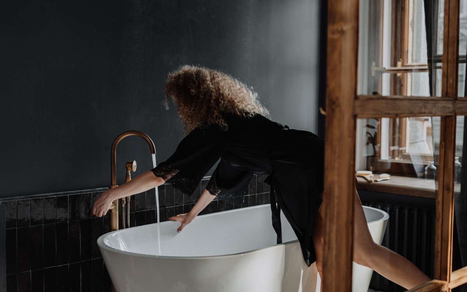 Una joven de pelo rizado prepara un baño en un elegante y moderno cuarto de baño con azulejos oscuros y luz natural.