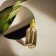 Una imagen minimalista que muestra una botella de suero cosmético transparente junto a una hoja verde en un espejo redondo.