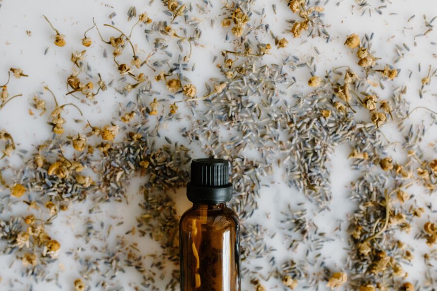Primer plano de una botella de aceite esencial rodeada de lavanda seca y manzanilla sobre una superficie blanca.