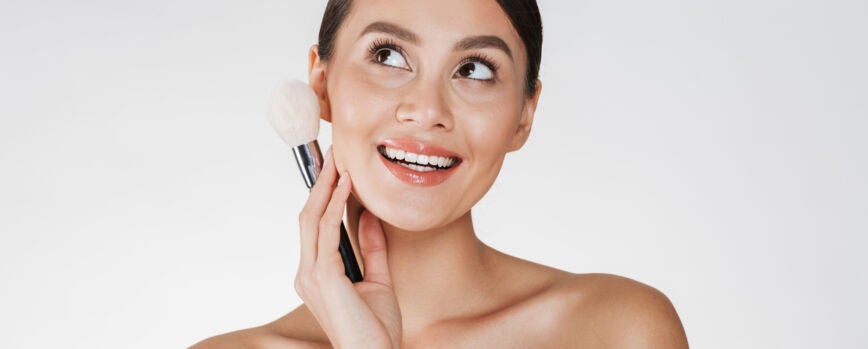 Foto de belleza de mujer bastante joven con el pelo en moño mirando hacia arriba y sosteniendo el pincel de maquillaje cerca de su rostro, aislado en blanco
