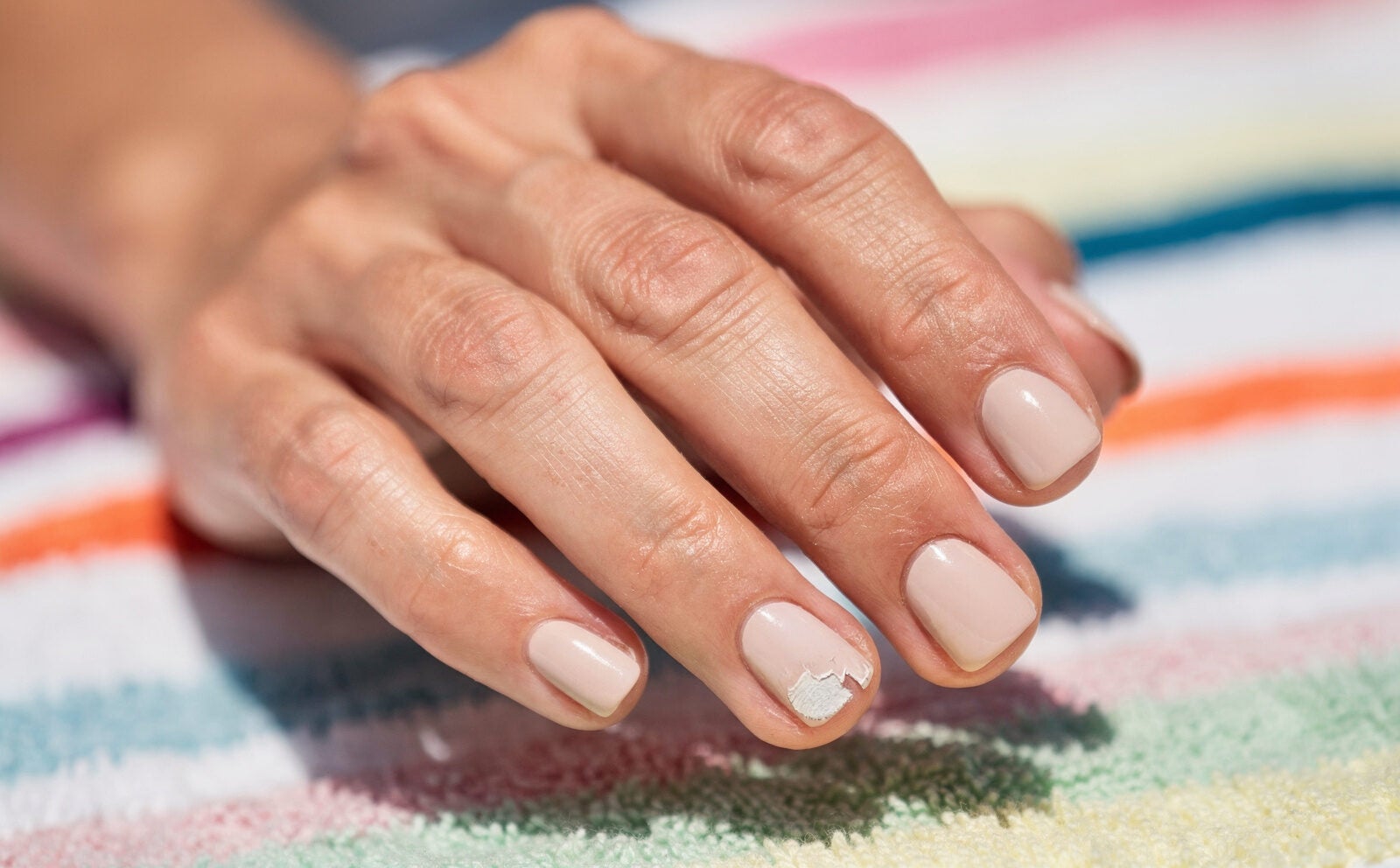 Mano con manicura cuadrada estilo años 90 con el esmalte ligeramente descascarillado en la punta de una uña, fondo de toalla de playa desenfocada, luz natural exterior. Sin manicura perfecta recién hecha.