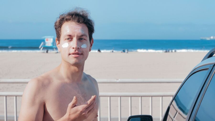Hombre sin camisa en la playa con crema solar en la cara, disfrutando de un día soleado.
