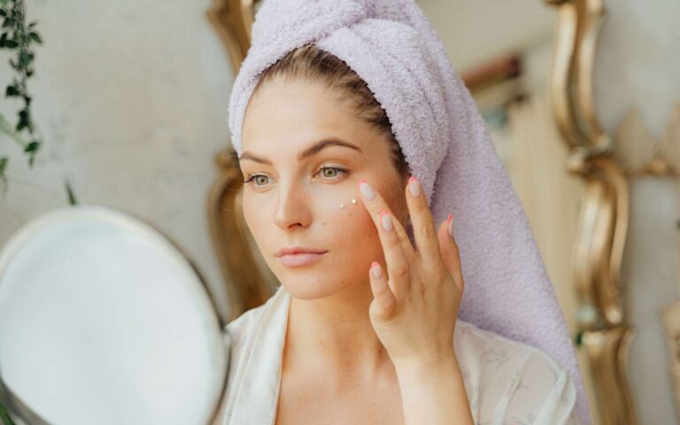 Mujer con toalla para la cabeza aplicando crema hidratante frente al espejo en el interior.