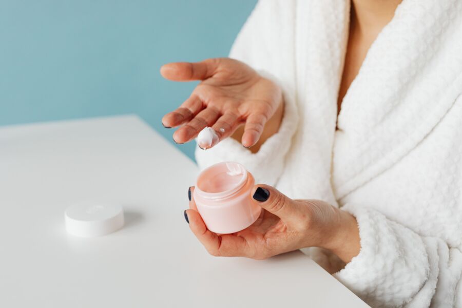 Una mujer en bata aplicando crema para el cuidado de la piel desde un frasco abierto, destacando la rutina hidratante.