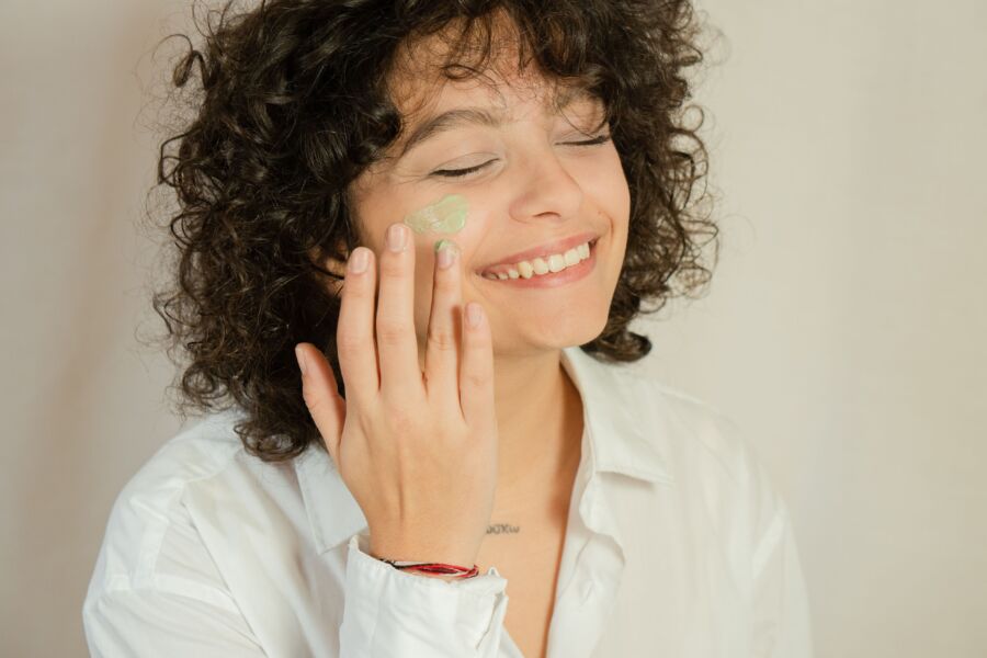Primer plano de una mujer sonriente aplicando crema facial con los ojos cerrados.