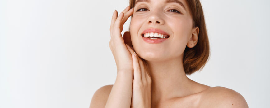 Belleza para el cuidado de la piel. Sonriente mujer natural con hombros desnudos y piel sana, limpia y fresca, luciendo feliz, tocando la mejilla. Chica aplicar cosméticos faciales, pared blanca
