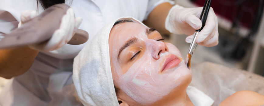 Esteticista con un pincel aplica una mascarilla hidratante blanca a la cara de un cliente joven en un salón de belleza spa