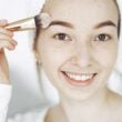 Retrato de una joven sonriente aplicando maquillaje con un pincel en interiores.