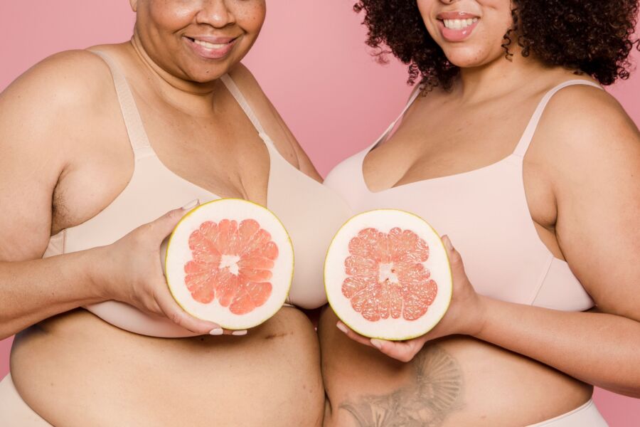 Modelos femeninos afroamericanos anónimos de talla grande en lencería con mitades de pomelo maduro en las manos sonriendo sobre fondo rosa en el estudio