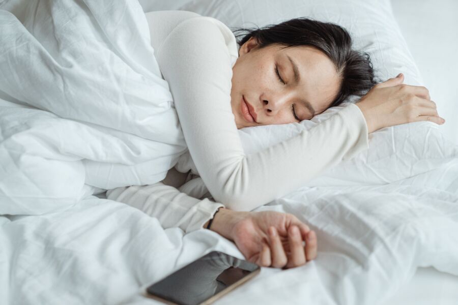 Mujer asiática tranquila con pijama blanco durmiendo en una cama cómoda con sábanas blancas cerca de un teléfono móvil moderno por la mañana