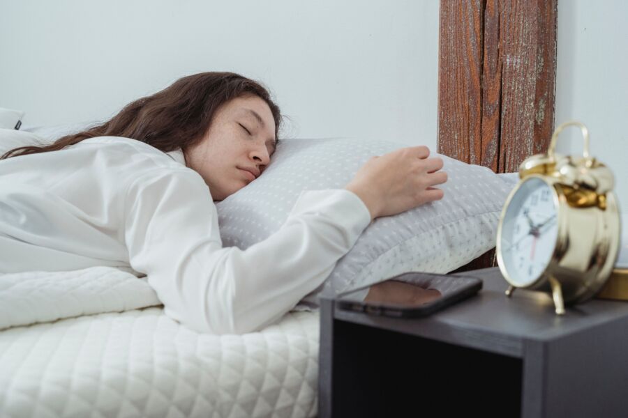 Mujer joven con cabello largo y ondulado oscuro durmiendo tranquilamente sobre el vientre en una cómoda cama bajo una manta blanca cerca de la mesita de noche con despertador y teléfono inteligente