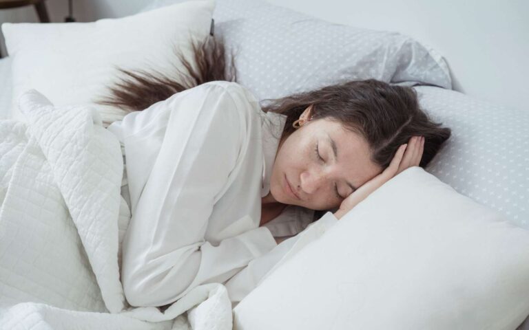 Una mujer serena descansa cómodamente en la cama con almohadas y mantas suaves, abrazando la relajación.