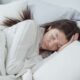 Una mujer serena descansa cómodamente en la cama con almohadas y mantas suaves, abrazando la relajación.