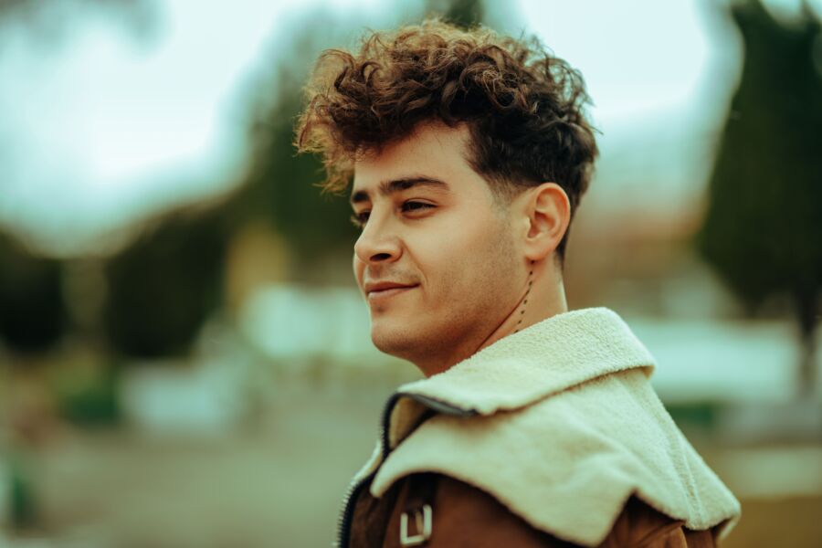 Joven de pelo rizado con chaqueta marrón disfrutando de un día soleado al aire libre.