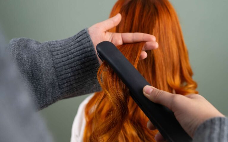 Manos peinando el pelo largo de color rojo con una plancha plana en interiores. Concepto de belleza y cuidado del cabello.